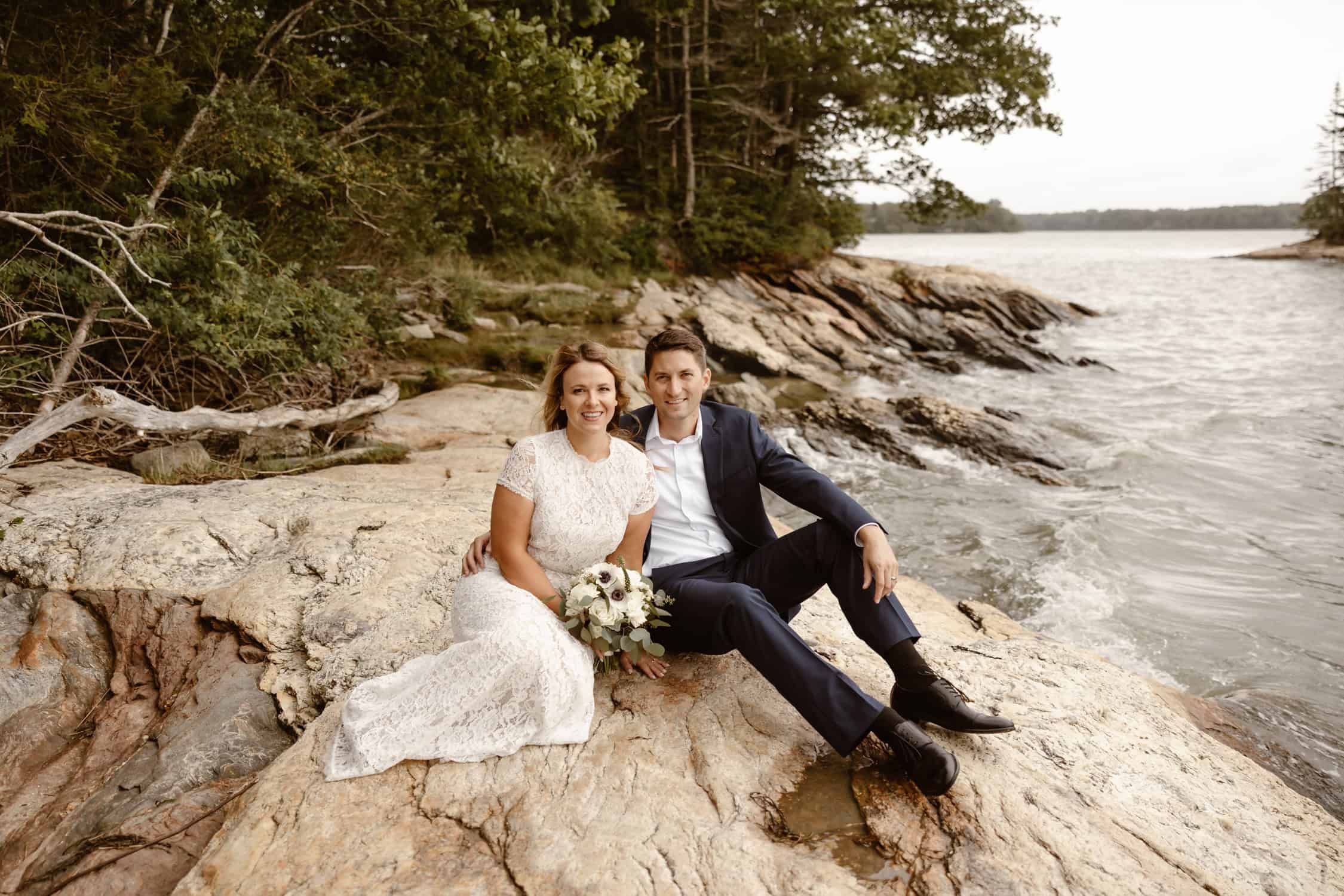 A couple sitting side by side on a rock by the ocean in Freeport, Maine. They are wearing their elopement attire, having just tied the knot!