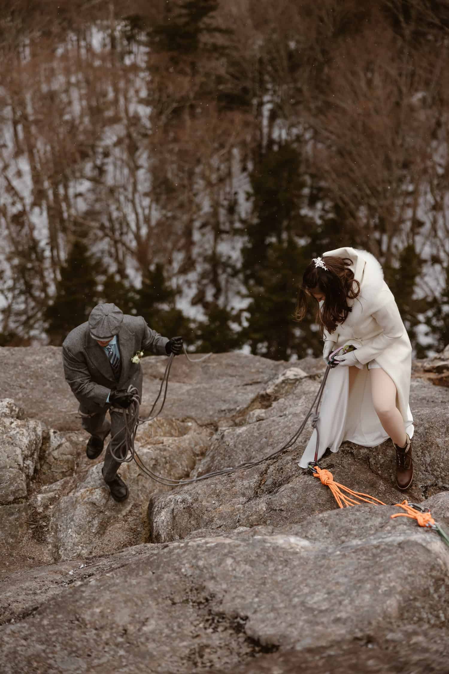 A bride and groom clipping in, preparing to rappel down the rock face, wearing their elopement attire.