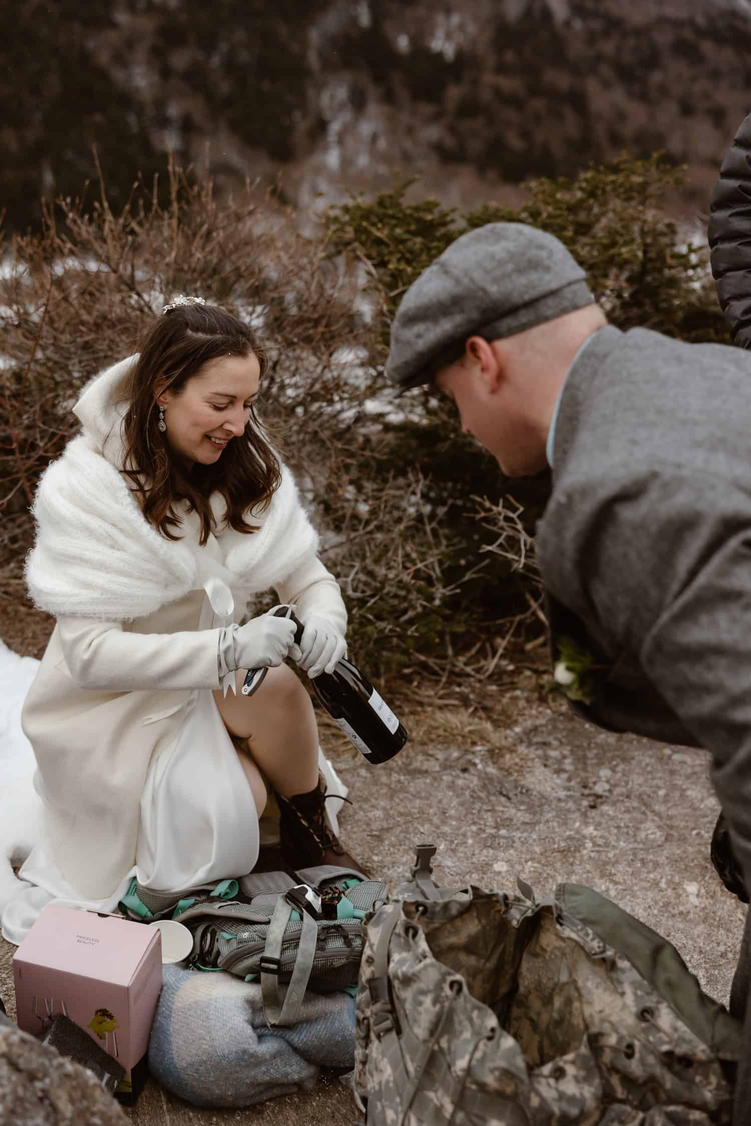 A bride opening a bottle of champagne, celebrating eloping after rappelling in the White Mountains.